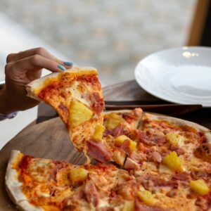 A close-up of a hand picking a slice from a freshly baked Hawaiian pizza on a wooden board.