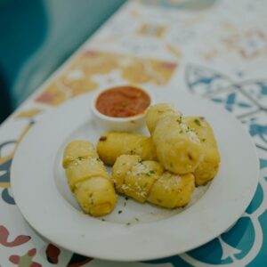 Close-up of golden-brown breadsticks topped with cheese, served with marinara on a vibrant patterned table.
