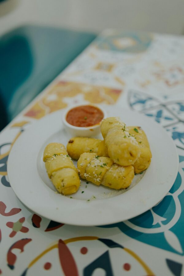Close-up of golden-brown breadsticks topped with cheese, served with marinara on a vibrant patterned table.