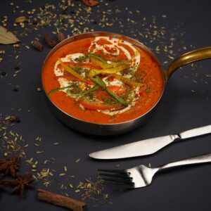 Close-up of a spicy red curry in a brass pot, garnished with herbs and surrounded by spices on a black background.